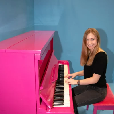 Junge Frau sitzt mit leichtem Lächeln an einem knallpinkfarbenen Klavier vor blauem Hintergrund.