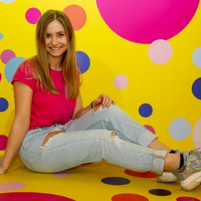 Lisa-Carolina sitzt lächelnd in lässiger Pose vor gelber Wand mit buntem, großem Punktmuster.