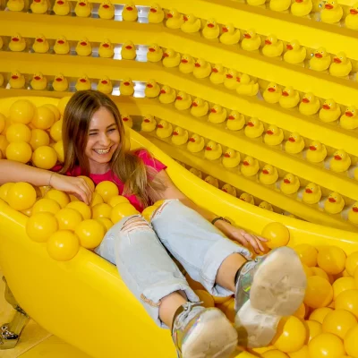 Frau sitzt lachend in einer mit gelben Bällen gefüllten Badewanne vor einer Wand voller gelber Quietscheentchen.