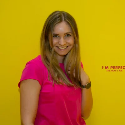 Frau mit langen blonden Haaren und pinkem T-Shirt vor gelber Wand mit dem Schriftzug „I'M PERFECT THE WAY I AM“