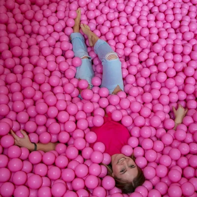 Junges weibliches Model liegt in rosa Bällebad, trägt pinkes Oberteil und hellblaue Jeans, Blick zur Kamera gerichtet.