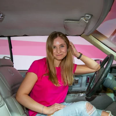Frau mit langen, offenen Haaren und pinkem T-Shirt sitzt auf dem Fahrersitz eines klassischen Cadillacs mit hellem Lederinterieur.
