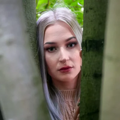 Porträtfotografie einer jungen Frau mit langen, hellblonden Haaren, die zwischen zwei Baumstämmen hervorschaut
