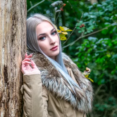 Junge Frau mit langen, hellblonden Haaren im beigen Mantel mit Fellkragen, die an einem Baum im grünen Wald lehnt