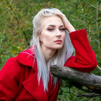 Weibliches Model mit langen, hellblonden Haaren und rotem Mantel lehnt an einem Baumstamm im Wald.