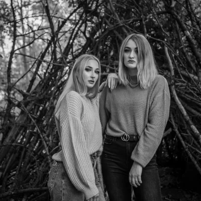 Schwarz-weiße Porträtfotografie von zwei jungen Frauen mit langen Haaren im Pullover vor einer Kulisse aus Ästen im Wald