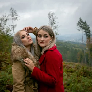 Zwei junge Frauen mit langen, blonden Haaren stehen auf einer Lichtung, im roten Mantel bzw. im beigen Mantel mit Fellkragen.