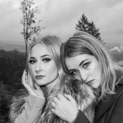 Zwei junge Frauen mit langen Haaren, eng aneinander gelehnt, im Mantel mit Fellkragen, schwarz-weiß, vor Waldlandschaft