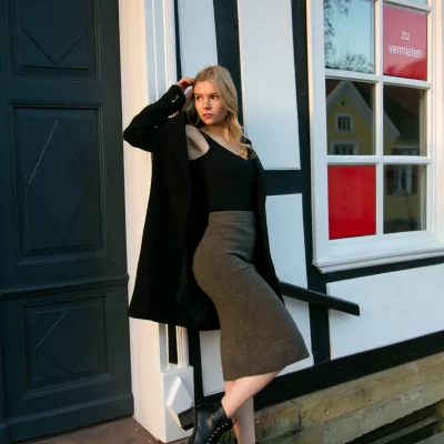 Frau in schwarzer Jacke und grauem Rock lehnt an einer Hauswand mit Fachwerk vor einer Eingangstür und einem Fenster.