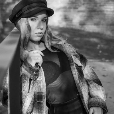Porträtfotografie eines weiblichen Models mit Schiebermütze und kariertem Mantel, das sich an ein Metallgeländer lehnt