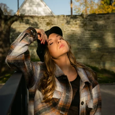 Frau mit geschlossenen Augen, langem blondem Haar und kariertem Mantel, die im Sonnenlicht vor einer Steinmauer steht
