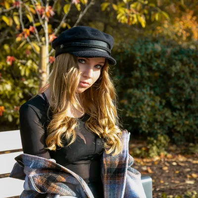 Frau mit langen blonden Haaren und rosafarbener Mütze, die entspannt an einem Schaufenster lehnt und direkt in die Kamera blickt.