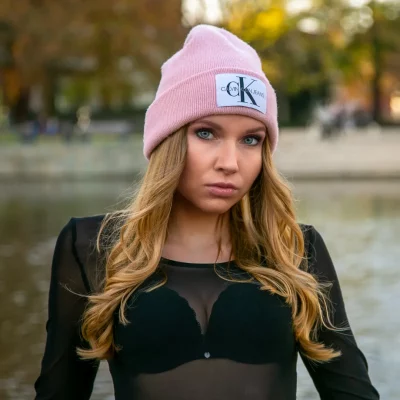 Frau mit langen blonden Haaren, pinker Mütze und schwarzem, transparentem Oberteil vor unscharfem Hintergrund mit Wasser