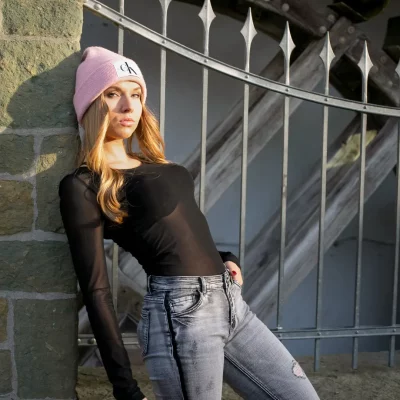 Frau mit langen, blonden Haaren, rosafarbener Mütze und schwarzem Oberteil und Jeans vor einem historischen Mühlrad