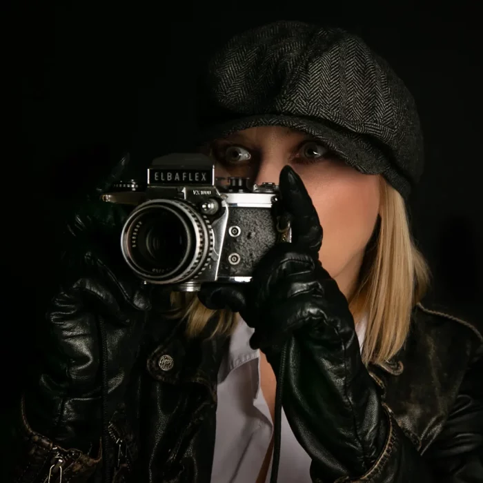 Weibliches Model mit Schirmmütze und Handschuhen hält eine Elbaflex-Kamera im Detektiv-Stil vor dunklem Hintergrund.