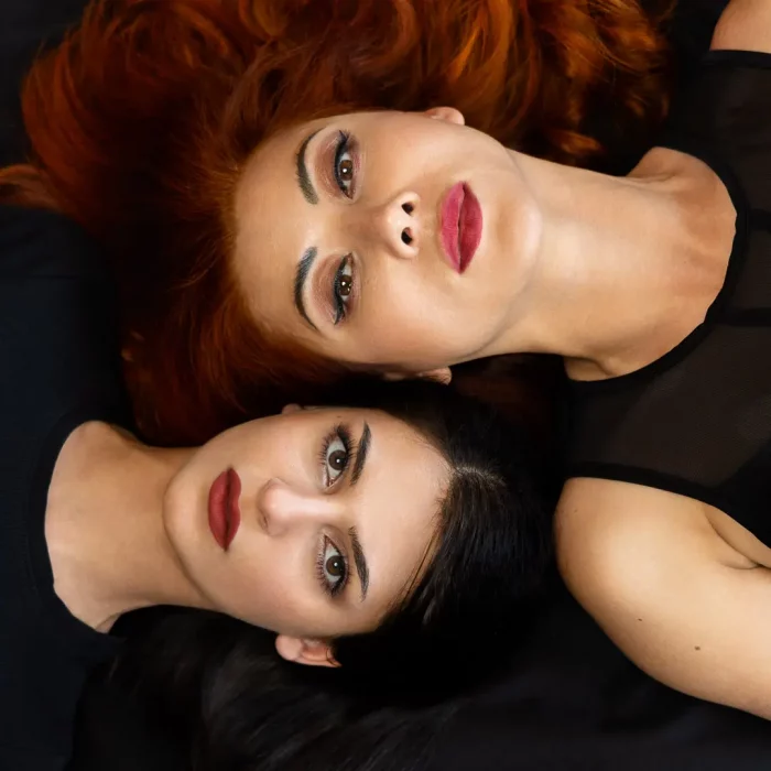Zwei Frauen mit roten und dunklen Haaren, die Rücken an Rücken auf schwarzem Untergrund liegen und in die Kamera blicken.