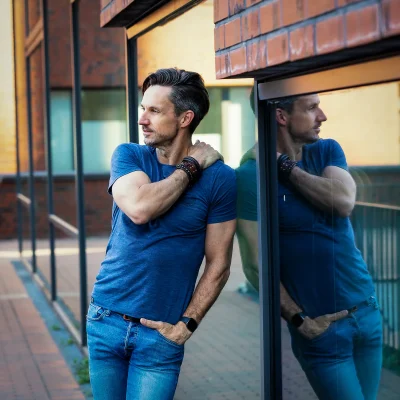 Mann mit Bart in blauer Jeans und blauem T-Shirt, der seitlich an großem Fenster einer Backsteinfassade lehnt