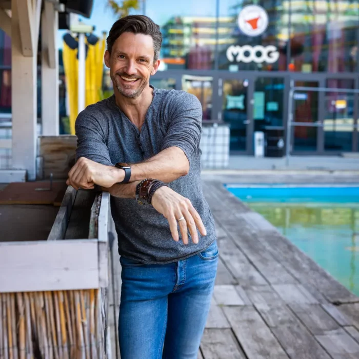 Lächelnder Mann in legerer Kleidung an einer Poolbar im Außenbereich mit Holzplanken und dem Schriftzug „MOOG“ im Hintergrund