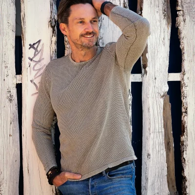 Portraitfotografie eines lächelnden Mannes mit Bart, der vor einer rustikalen Holzwand in Jeans und hellem Pullover steht