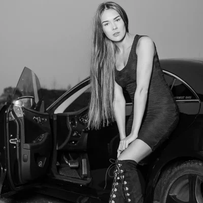 Auto-Fotoshooting Soest in NRW: Frau mit langem Haar in engem Kleid und hohen Stiefeln lehnt an offenem Türrahmen eines Autos.