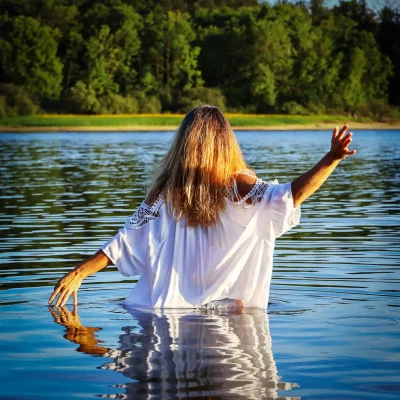 Frau mit langen Haaren im weißen Oberteil steht hüfttief im Wasser und hebt einen Arm