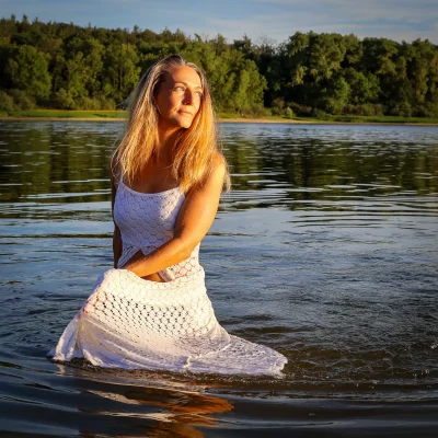 Frau im weißen Kleid kniet im Wasser eines Sees vor bewaldetem Ufer und blickt nach rechts oben