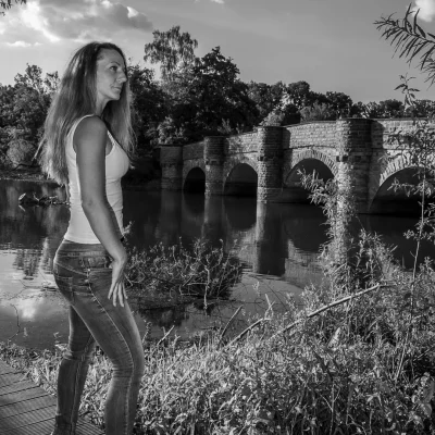 Porträtfotografie einer Frau in Jeans und Top, stehend am Flussufer vor einer historischen Steinbogenbrücke bei Tageslicht