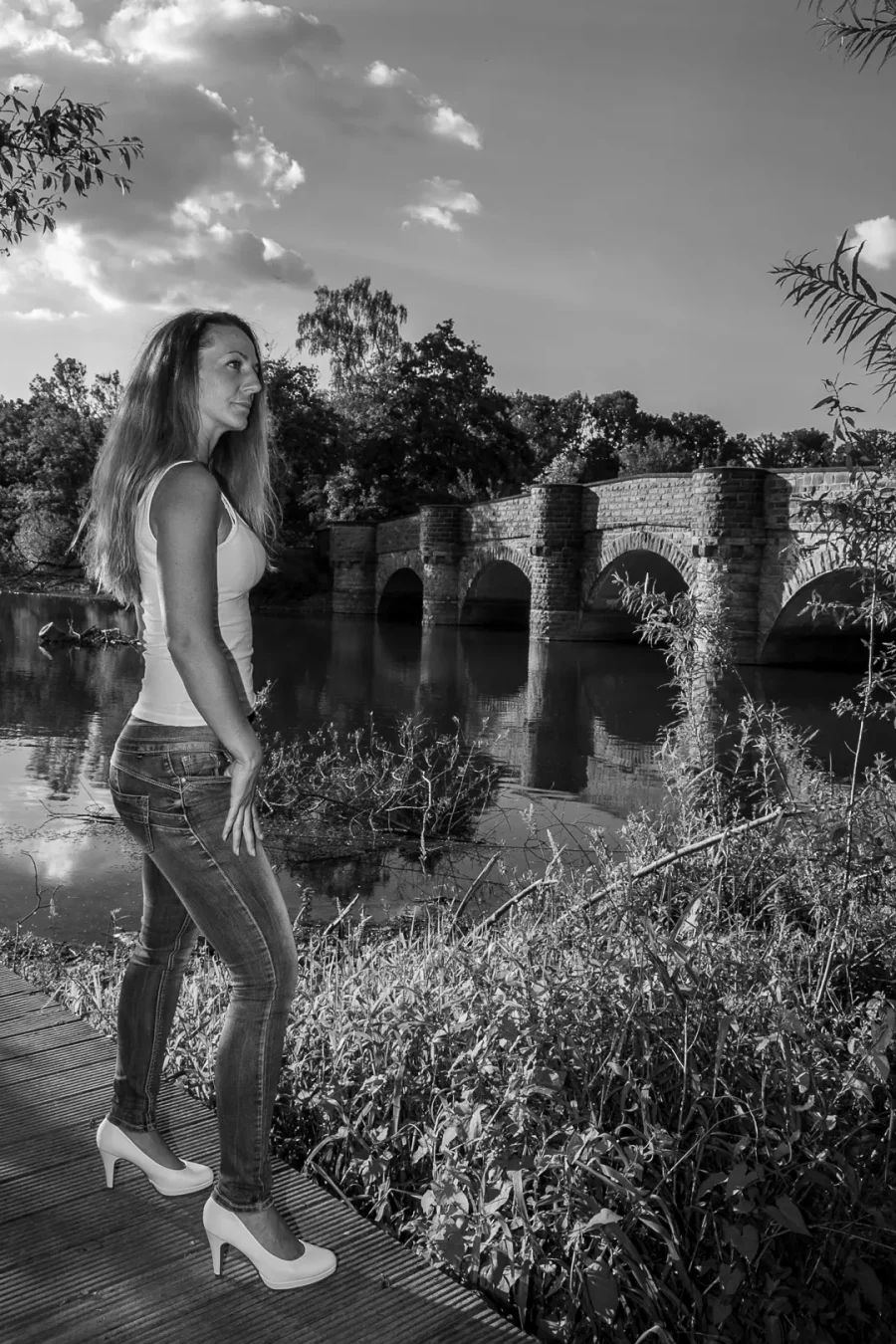 Porträtfotografie einer Frau in Jeans und Top, stehend am Flussufer vor einer historischen Steinbogenbrücke bei Tageslicht