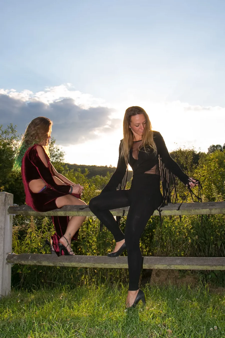 Zwei Frauen in auffälliger Kleidung auf Holzzaun, Sonnenuntergang im Hintergrund, grüne Natur, sommerliche Atmosphäre