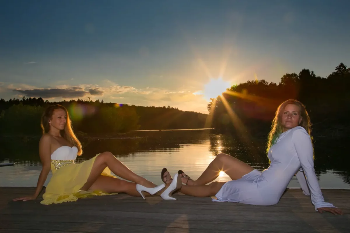 Zwei Frauen sitzen auf einem hölzernen Steg am See, Sonnenuntergang im Hintergrund.