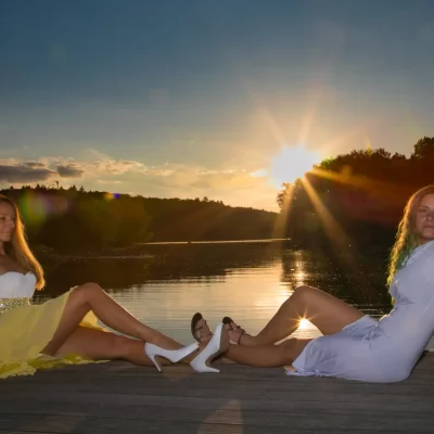 Zwei Frauen sitzen auf einem hölzernen Steg am See, Sonnenuntergang im Hintergrund.