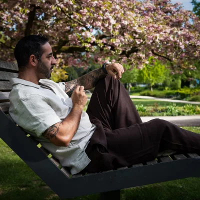 TFP-Porträt von Nevio Montefusco, zurückgelehnt auf einer Parkbank vor rosa Kirschblüten, Leinenhemd und Sonnenbrille in der Hand.