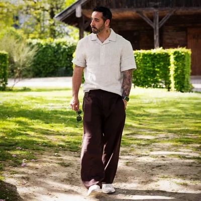 Nevio geht über eine Wiese vor einer Holzhütte, helles Leinenhemd und weite braune Hose, Sonnenbrille in der Hand – Available-Light-Outdoor.