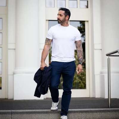 Nevio in weißem T-Shirt und dunkelblauer Hose auf Treppenstufen, Nadelstreifenjacke über dem Arm, vor heller Fassade.