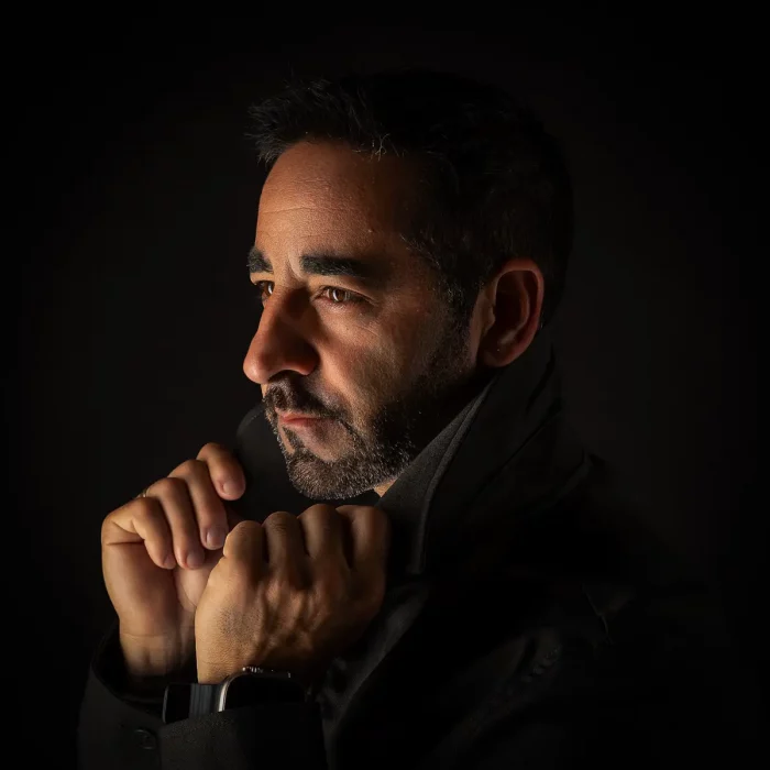 Lowkey-Profilportrait von Nevio im dunklen Mantel, dramatisches Seitenlicht vor schwarzem Hintergrund.