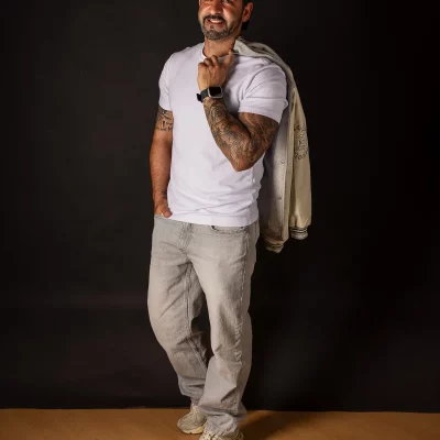 Ganzkörperaufnahme aus dem TFP-Studioshooting: Nevio in weißem T-Shirt, heller Jeans und College-Jacke über der Schulter.