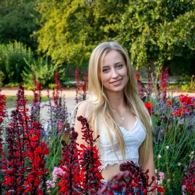 Porträtfotografie einer jungen Frau mit langen blonden Haaren, die zwischen roten Blüten im Garten steht und freundlich lächelt.
