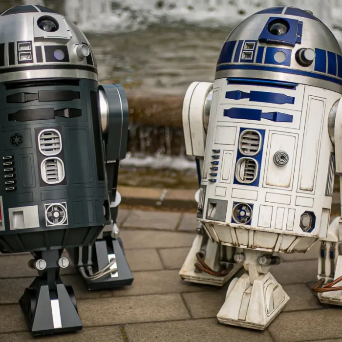 Zwei R2D2-Roboter-Modelle in Originalgröße stehen vor einem steinernen Brunnen auf gepflastertem Boden.