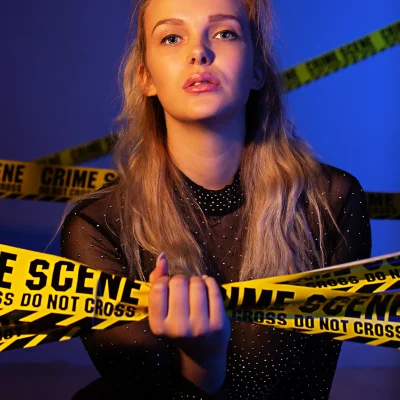Portraitfotografie eines weiblichen Models mit langen, offenen Haaren, das ein gelbes Crime-Scene-Absperrband vor blauem Hintergrund hält