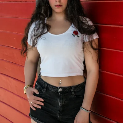 Weibliches Model mit langem, lockigem Haar in weißem, kurzem Shirt und schwarzen Jeansshorts lehnt an roter Holzwand in Soest, großes Bein-Tattoo