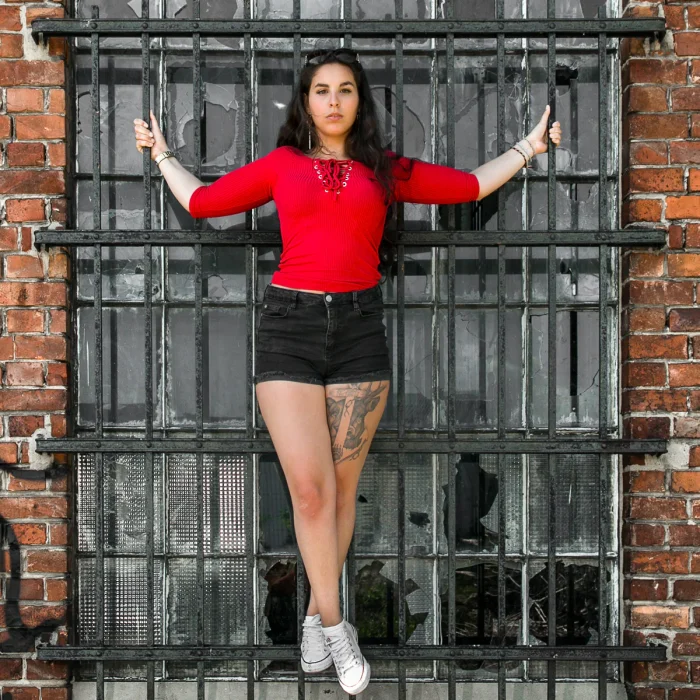 Lost Place Fotografie NRW: Sarah mit rotem Oberteil, Bein-Tattoo und schwarzen Haaren steht mit ausgestreckten Armen auf einem eisernen Fenstergitter.
