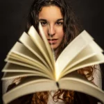 Kreatives Fotoshooting: Junge Frau mit langen, lockigen Haaren, die ein aufgeschlagenes Buch in den Händen hält und direkt in die Kamera blickt.