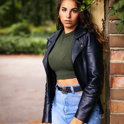 Junge Frau mit langen dunklen Haaren, grünem Oberteil, schwarzer Lederjacke und Jeans, die an einer gemauerten Torwand lehnt