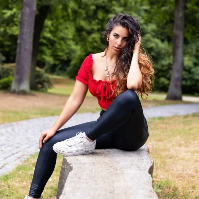 Frau mit langen, dunklen Haaren, rotem Oberteil und schwarzen Leggings sitzt auf einem Stein in einem Park in Hamm.