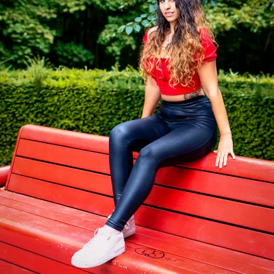 Frau mit langen, lockigen Haaren, rotem Oberteil und glänzender, dunkler Hose sitzt seitlich auf der Rückenlehne einer großen roten Parkbank.