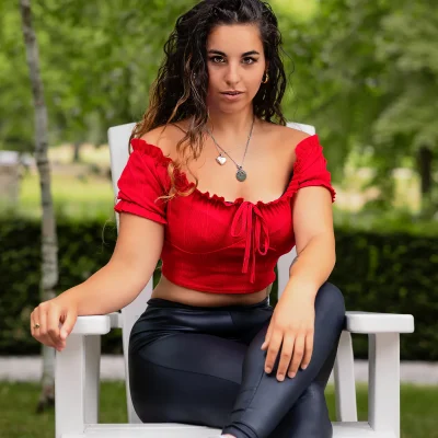 Model Sarah Ben Brahim mit lockigen Haaren in rotem Crop-Oberteil und schwarzen Leggings auf weißem Stuhl im Freien