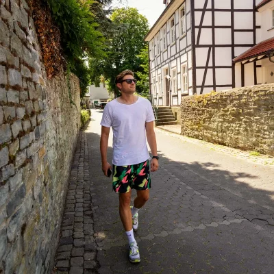 Stefan mit Sonnenbrille und weißem T-Shirt läuft durch eine schmale Altstadtgasse mit Fachwerkhaus und Natursteinmauer.