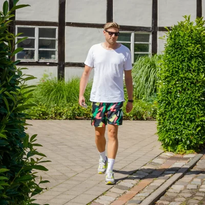 Männliches Model in weißem T-Shirt und gemusterten Shorts läuft zwischen zwei grünen Hecken auf einem gepflasterten Weg.