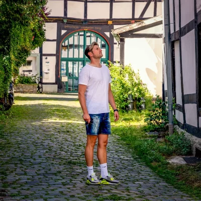 Männliches Model im weißen T-Shirt und Jeansshorts steht auf Kopfsteinpflaster vor einem historischen Fachwerkhaus, Sommerlicht