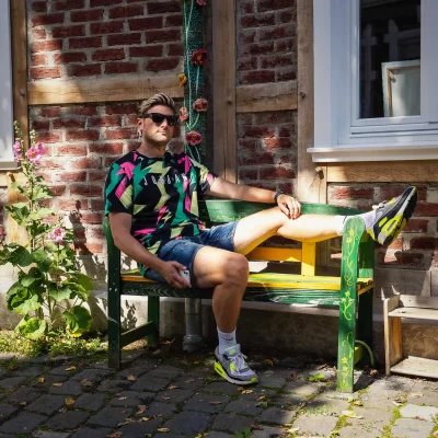 Porträtfotografie eines jungen Mannes mit Sonnenbrille und bunt gemustertem T-Shirt, lässig auf grüner Bank vor Backsteinwand sitzend